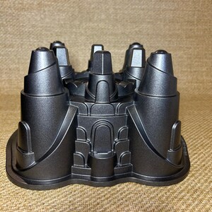 May include: A dark gray, castle-shaped cake pan. The pan features detailed turrets, arched doorways, and a textured surface. The baking pan is designed to create a castle-themed cake, perfect for themed parties or special occasions.