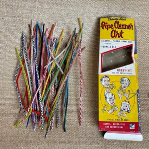 May include: A collection of colorful pipe cleaners in various colors and patterns, next to a vintage box labeled "Pipe Cleaner Art." The box is yellow with illustrations and text, advertising it as a hobby kit for all ages.
