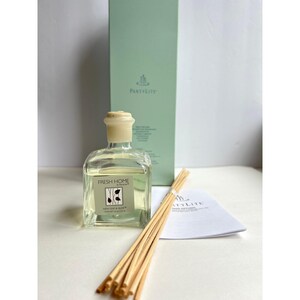May include: A clear glass bottle with a beige cap and a light green liquid inside. The bottle is labeled "FRESH HOME" and "NEW LINEN & ALOE". A bundle of wooden sticks and a green box with the "Partylite" logo are also present.