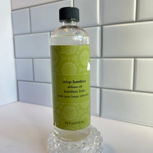 May include: A clear glass bottle of "crisp bamboo" diffuser oil with a black cap. The label is green with a floral pattern and text in English and French. The bottle contains 16 fl oz / 473 mL of oil.