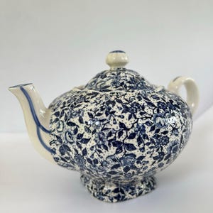 May include: A ceramic teapot with a blue floral pattern on a white background. The teapot has a curved spout, a handle, and a lid with a small knob. The design features small blue flowers and dots, creating a classic look.
