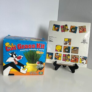 May include: A blue Cat Grass Kit box featuring Sylvester and Tweety, with a pot of growing grass. Next to it is a vintage Looney Tunes art print with various cartoon characters.