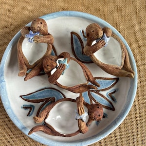 May include: A ceramic plate with a blue and white floral design. Three brown monkey figurines are attached to the plate, each holding a blue starfish. The plate has a blue rim and is set against a neutral background.