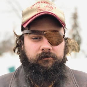 May include: A man with a long beard and a leather eye patch is wearing a brown leather jacket, a yellow button-down shirt, and a baseball cap that says "Rottz's Livestock 717-532-5891".