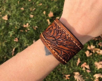 May include: A brown leather cuff bracelet with a floral design and tan stitching. The bracelet is worn on a person's wrist.