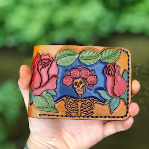 May include: A brown leather wallet with a Day of the Dead design. It features a skeleton with a floral crown, framed by red roses and green leaves. The background is blue.