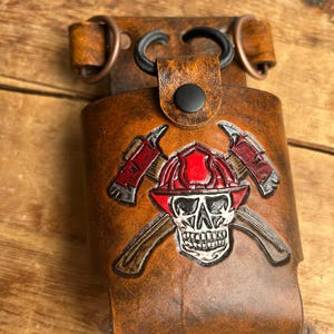 Peut inclure: Porte-radio en cuir marron avec un motif de crâne de pompier et de haches croisées. Le support est doté d'une boucle pour l'attacher à une ceinture ou à une sangle. Le motif présente des accents rouges, blancs et noirs.