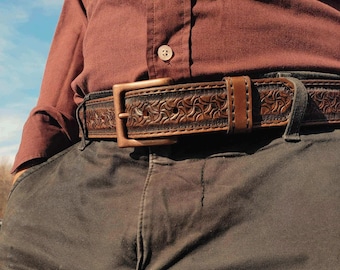Puede incluir: Un cinturón de cuero marrón con un patrón floral decorativo y una hebilla de color cobre. El cinturón se lleva con pantalones grises oscuros y una camisa burdeos abotonada. El cinturón mide aproximadamente 3,8 cm de ancho.