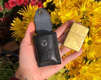 May include: A black leather lighter case with a decorative J on the front. The case is open and a gold lighter is visible inside. The case is held in a hand against a background of yellow and red flowers.