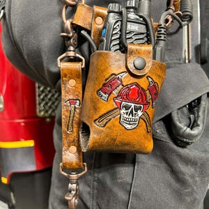 May include: A brown leather radio holder with a firefighter skull and crossed axes design. The holder is attached to a harness with a radio and a strap with a small axe. The leather has a distressed look.