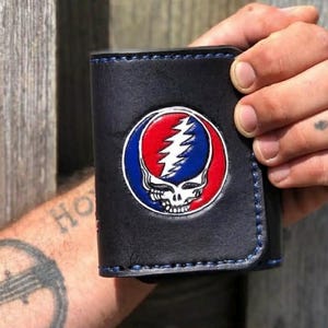 May include: Black leather wallet with a colorful Grateful Dead Steal Your Face logo. The wallet has blue stitching around the edges. The logo features a skull with a lightning bolt, surrounded by a red, white, and blue circle.