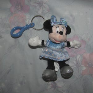 Könnte beinhalten: Ein Minnie Mouse Plüsch-Schlüsselanhänger. Das Plüschtier trägt ein hellblaues Kleid und eine Schleife mit silbernen Akzenten. Es hat schwarze Ohren, schwarze Beine, weiße Handschuhe und silberne Schuhe. Ein blauer Kunststoffclip ist am Schlüsselanhänger befestigt.