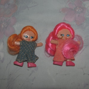 May include: Two small vintage dolls with long, colourful hair. One doll has orange hair and a blue patterned dress, the other has pink hair and a pink dress. Both dolls have pink shoes and blue eyes.