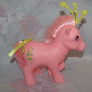 May include: A pink toy pony with a pink mane and tail, adorned with a yellow ribbon and star-shaped accents. The pony has green and yellow star graphics on its side and a yellow star crown. The toy is a collectible.