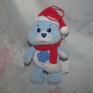 May include: A light blue plush bear wearing a red Santa hat and scarf. The bear has a blue cloud design on its belly and a heart-shaped nose. The scarf has white trim. A small tag is attached to the bear's foot.