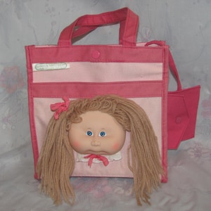 May include: A pink and white tote bag featuring a doll face with blonde yarn hair, blue eyes, and a pink bow. The bag has pink handles and a side pocket. The text "Cabbage Patch Kids" is visible on the bag.