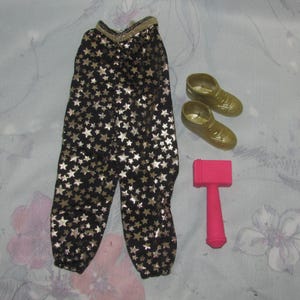 Könnte beinhalten: Ein Puppenoutfit mit schwarzer Hose mit goldenen Sternmustern, goldenen Schuhen und einem rosa Hammer. Die Hose hat einen goldenen Bund und ist an den Knöcheln gerafft. Die Schuhe sind metallisch goldfarben.