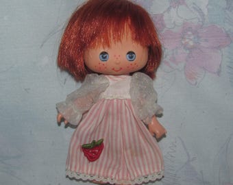 May include: A vintage doll with red hair, blue eyes, and freckles. The doll wears a pink and white striped dress with a strawberry applique and white lace trim. The doll's sleeves are sheer and ruffled.