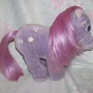 May include: A vintage lavender plush toy pony with a pink mane and tail. The pony has three gold flower accents on its side and large purple eyes. The toy is in a standing position.