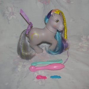 May include: A vintage lavender toy pony with a rainbow mane and tail, a yellow crescent moon on its flank, and accessories including two brushes, a pink bow, and a blue cloud. The pony has a purple ribbon tied to its tail.