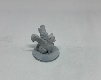 May include: A small, gray figurine of a winged cat, also known as a cat-gryphon, sits on a circular base. The cat has detailed wings and a curious expression. The figurine is made of a solid, matte material.