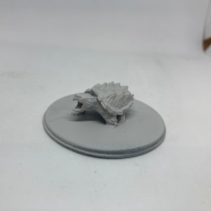 Könnte beinhalten: Eine kleine, graue, 3D-gedruckte Figur eines Schnappschildkröten-Alligators mit offenem Maul. Die Figur steht auf einem runden, grauen Sockel.