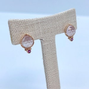 May include: A pair of rose quartz earrings with a pink gemstone accent. The earrings are displayed on a beige fabric stand.