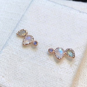 May include: A pair of earrings with three small, teardrop-shaped gemstones in each earring. The gemstones are iridescent and have a white, blue, and green color. The earrings are made of gold.