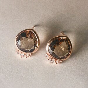 May include: A pair of gold earrings with a teardrop-shaped brown gemstone and a row of small clear stones around the bottom of each earring.