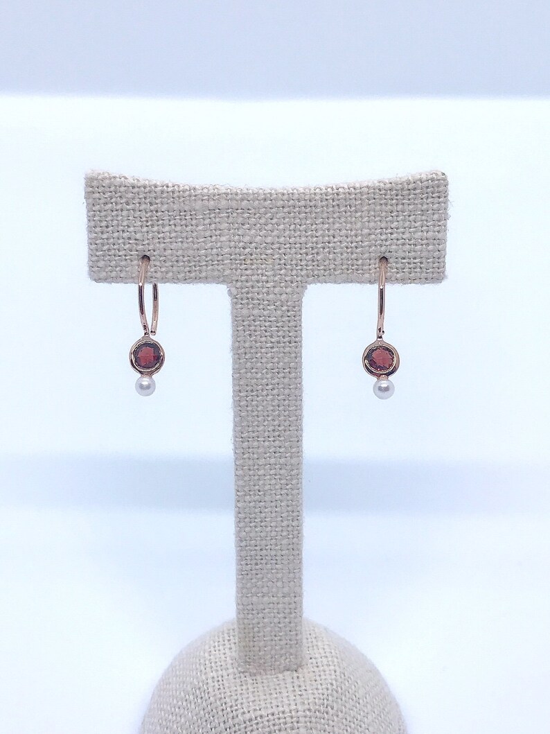 May include: A pair of rose gold hoop earrings with a small red gemstone and a pearl dangling from each hoop.