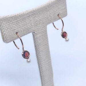 May include: A pair of rose gold hoop earrings with a red gemstone and a white pearl dangling from each hoop.