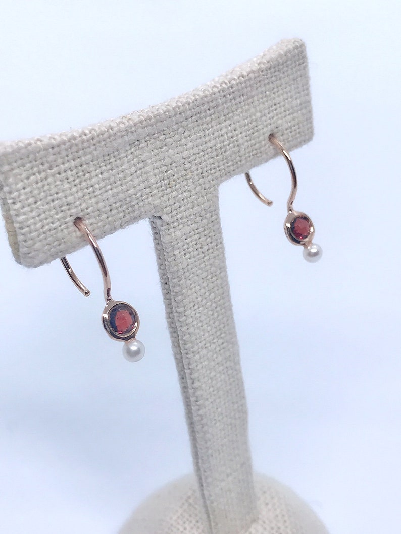 May include: A pair of rose gold hoop earrings with a red gemstone and a white pearl dangling from each hoop.