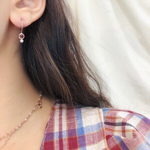 May include: A close-up of a person's ear wearing a delicate gold hoop earring with a small pink gemstone and a pearl dangling below.