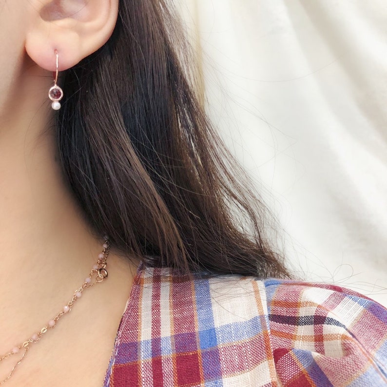 May include: A close-up of a person's ear wearing a delicate gold hoop earring with a small pink gemstone and a pearl dangling below.