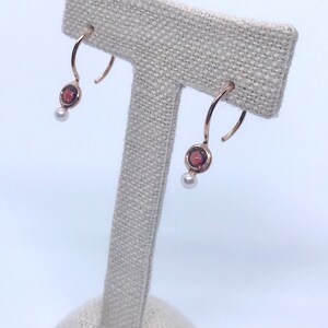 May include: A pair of gold hoop earrings with a small red gemstone and a pearl dangling from each hoop.