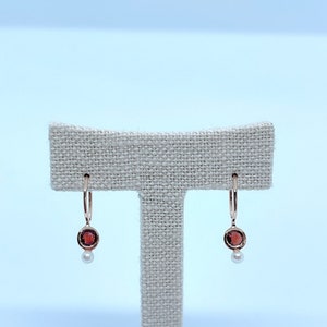 May include: A pair of gold hoop earrings with a small red gemstone and a pearl dangling from each hoop.