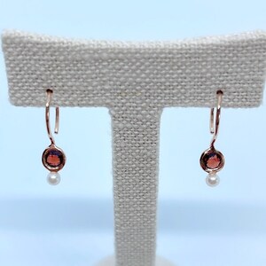 May include: A pair of rose gold earrings with a small round garnet stone and a pearl dangle.