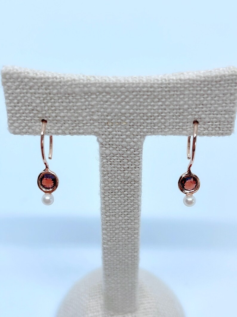 May include: A pair of rose gold earrings with a small round garnet stone and a pearl dangle.