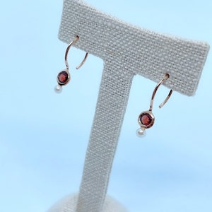 May include: A pair of gold hoop earrings with a small red gemstone and a pearl dangle. The earrings are displayed on a white fabric stand.