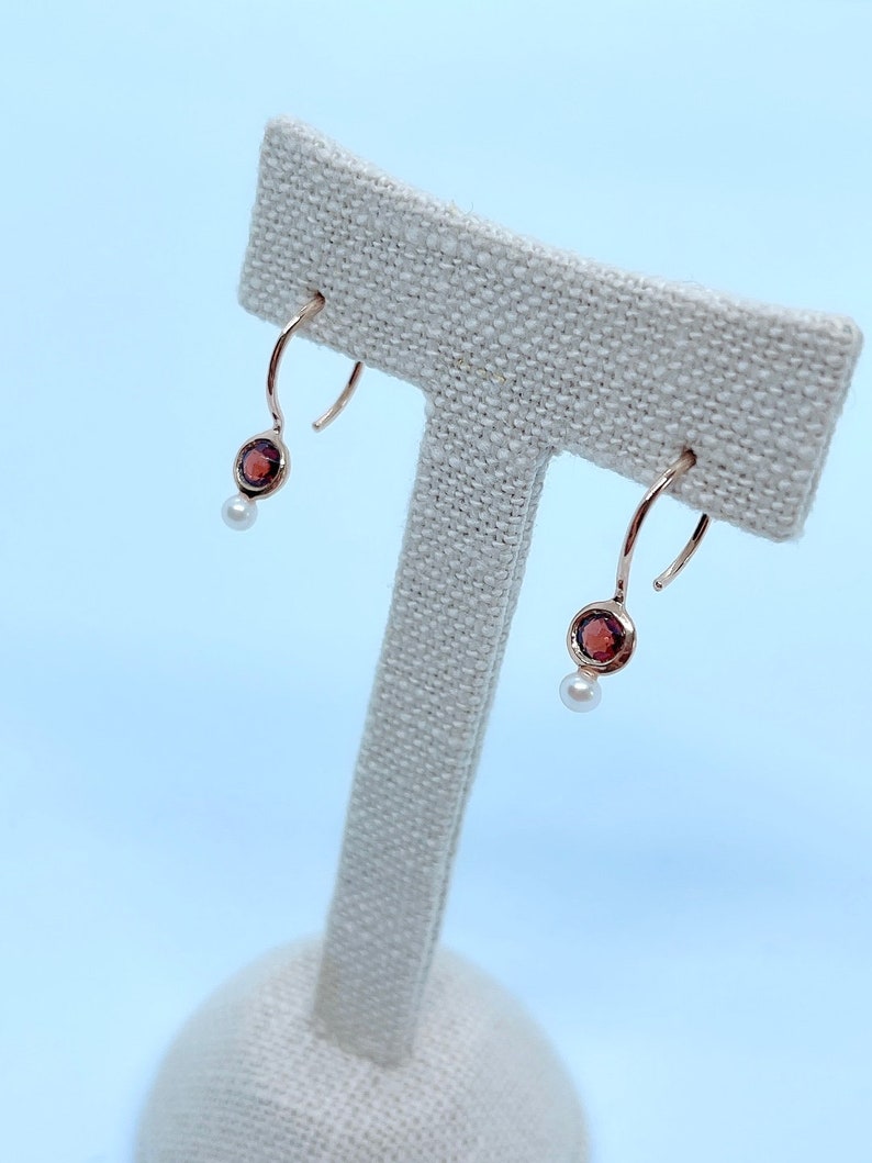 May include: A pair of gold hoop earrings with a small red gemstone and a pearl dangle. The earrings are displayed on a white fabric stand.