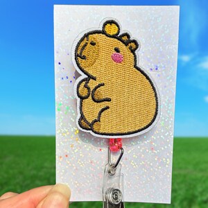 May include: A capybara-themed embroidered badge reel. The badge features a tan capybara with a pink cheek, a yellow crown, and black outlines. The badge is attached to a white card with a glittery effect, and a clear plastic reel with a pink bead.
