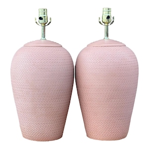May include: Two matching pink ceramic table lamps. Each lamp has a textured surface with a repeating wave pattern. The lamps have brass-colored hardware and a cylindrical neck. The lamps are a soft, muted pink color.