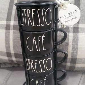 May include: A black stacking mug set with the words "ESPRESSO" and "CAFÉ" in white lettering. The mugs are held in a black metal stand. A white tag with the text "RAE DUNN by Magenta Stacking Mugs" is attached.