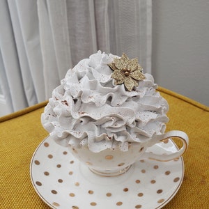 May include: A white teacup and saucer with gold polka dots. The teacup is filled with white whipped cream and topped with a gold glitter flower.