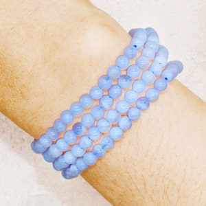 May include: Four light blue beaded bracelets are wrapped around a wrist. The bracelets are made of small, round beads. The skin tone is light. A white crystal is in the background.