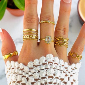 May include: A hand displaying a collection of gold rings. The rings feature various designs, including hearts, chain links, and a central oval-shaped stone. The rings are worn on multiple fingers, showcasing their different styles and sizes.