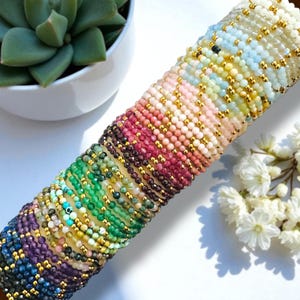 May include: A collection of beaded bracelets in a variety of colors, including shades of blue, green, pink, and purple. Each bracelet features small, round beads and gold-colored accents. The bracelets are arranged in a stacked display, showcasing the range of colors and designs.