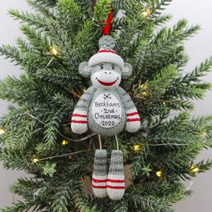 May include: A gray sock monkey Christmas ornament with red and white stripes. The ornament has the text "Beckham's 2nd Christmas 2022" on the front.