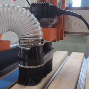 May include: A close-up of a black dust collection brush attached to a router, with a clear plastic hose connected to the brush. The router is mounted on a CNC machine, and the brush is positioned over a piece of wood.