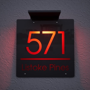 May include: A black address sign with the number "571" in red, illuminated lettering. The text "Listoke Pines" is below the number. The sign is mounted on a textured gray wall and has a black solar panel at the top.